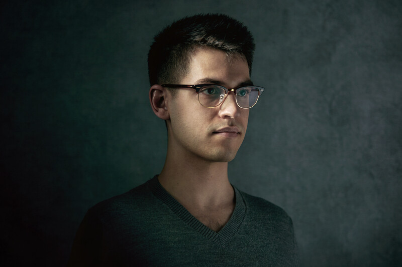 Young man wearing glasses and a dark green v-neck shirt against a teal studio backdrop.