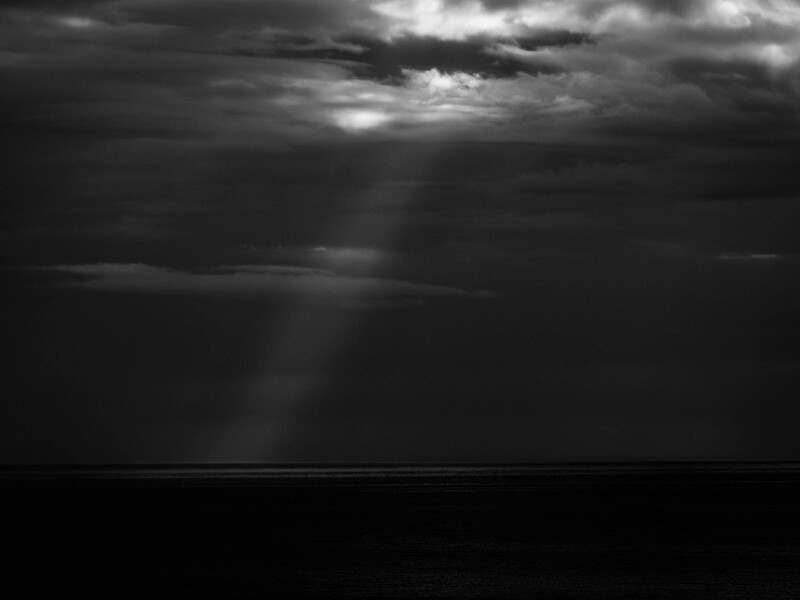 Dramatic black and white seascape with crepuscular rays breaking through storm clouds over a dark ocean horizon.