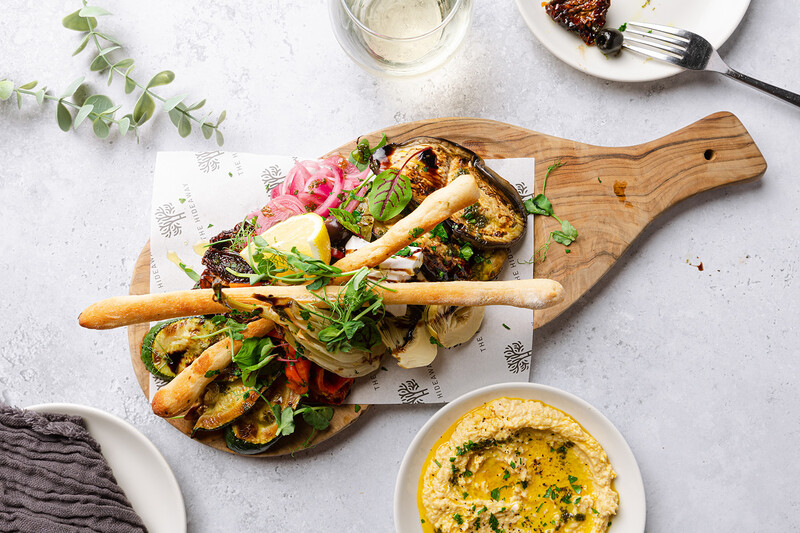 Grilled vegetables and breadsticks arranged on a wooden paddle board with hummus dip.