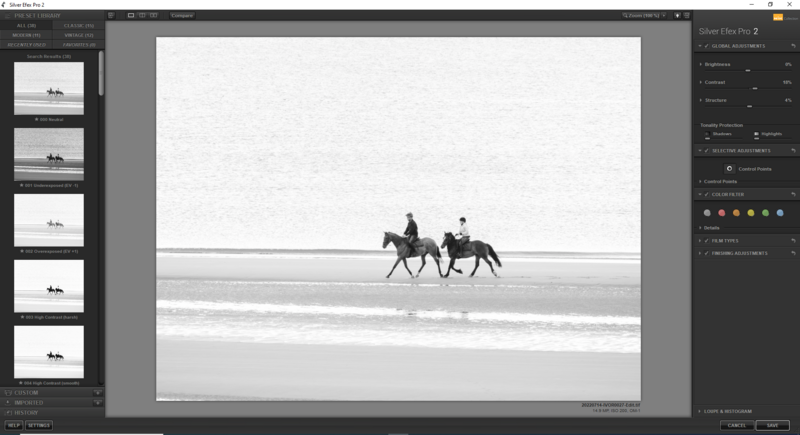 Two riders on horseback walking along a beach shoreline in black and white.
