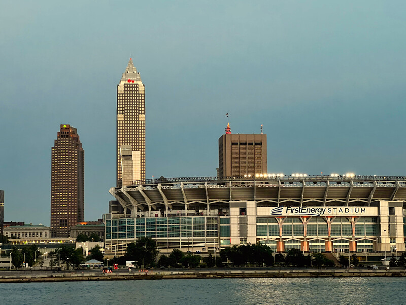 Waterfront cityscape with tall skyscraper, stadium, and historic buildings along a river at dusk.
