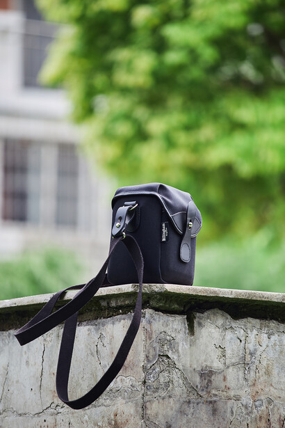 Black camera bag with shoulder strap positioned on concrete ledge with blurred green foliage in background.
