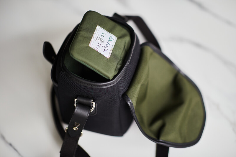 Black camera bag with olive-green interior and flap pocket containing a white label tag.