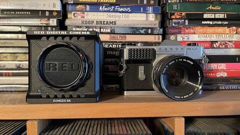 RED digital cinema camera body next to vintage lens on wooden shelf with DVDs stacked behind.