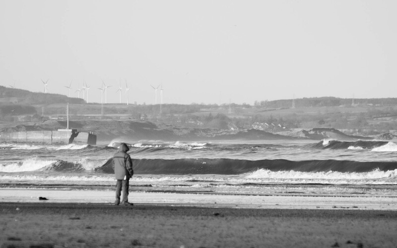 Surfer in wetsuit standing in shallow water holding surfboard, facing ocean waves.