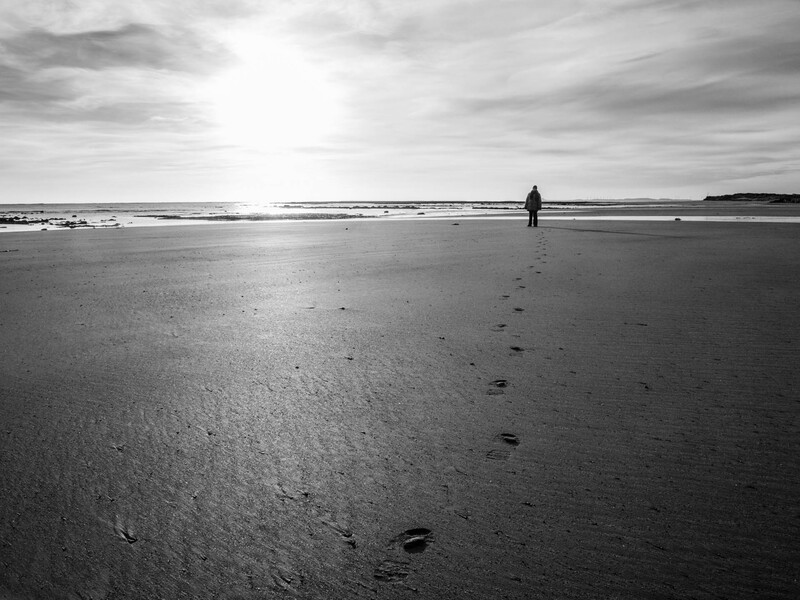 Solitary figure standing on a vast, wet beach with footprints trailing behind in the sand.