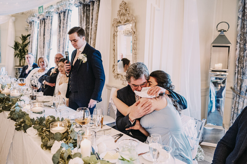 Man embracing woman at formal wedding reception table with guests in background.