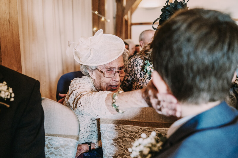 Elderly woman in white hat embracing bride during wedding ceremony reception.