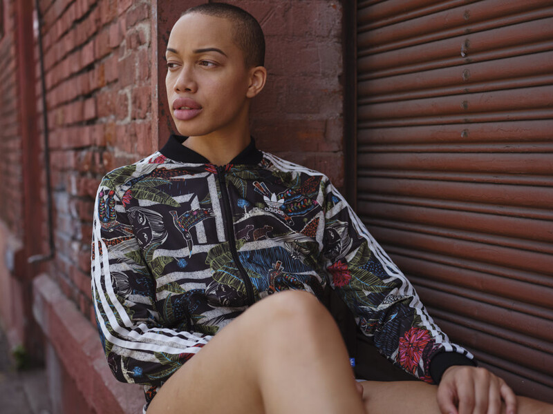 Person in patterned track jacket seated against brick wall with metal shutter background.
