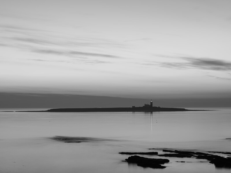 Minimalist black and white seascape with a distant lighthouse on the horizon and dark rocks in the shallow foreground.