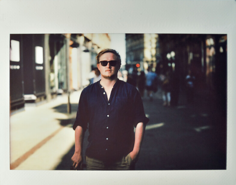Man in dark button-up shirt and sunglasses standing on an urban street with blurred pedestrians and storefronts in background.