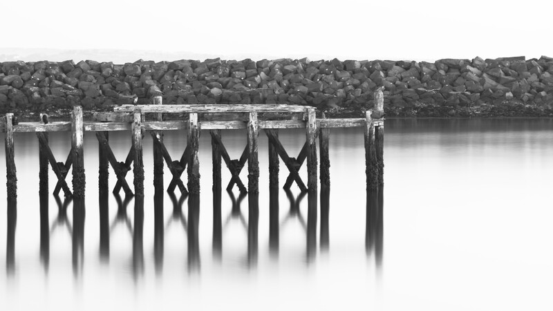 Wooden pier pilings with X-braced cross-beams extending into calm water, rendered in black and white.