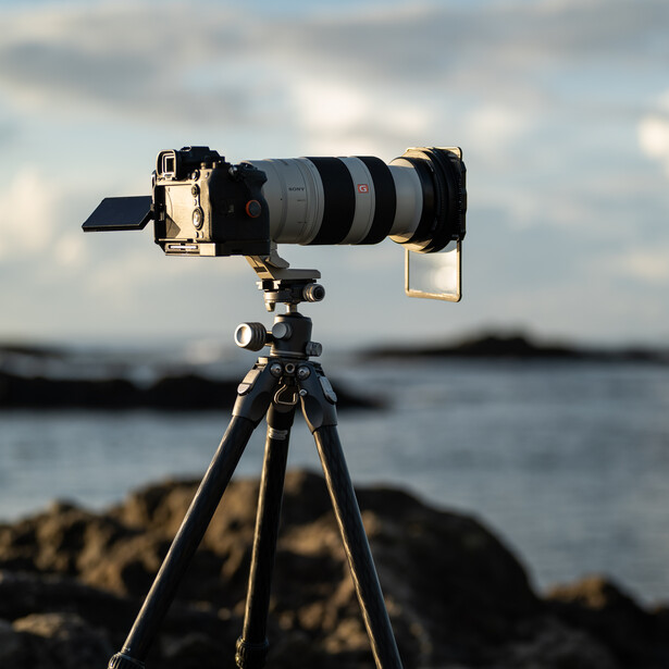 Professional telephoto lens mounted on tripod with camera body overlooking coastal rocks and water.