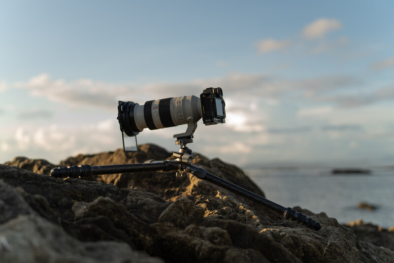 Telephoto lens mounted on tripod atop rocky coastal outcrop with calm water and distant landmass visible.