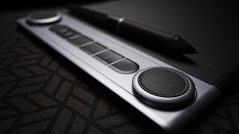 Graphics tablet with stylus pen and control dials on dark textured surface.