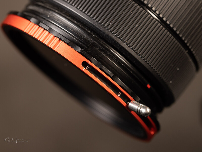 Close-up of a camera lens with orange and black rubber focus ring against neutral background.