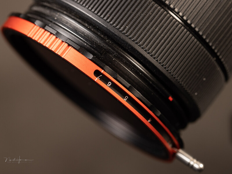Close-up of a camera lens with orange and black focus ring against neutral background.