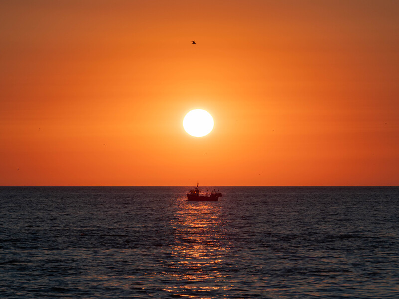 Fishing boat silhouetted against a brilliant orange sunset over calm ocean water.