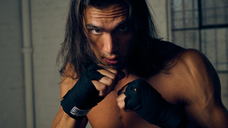 Shirtless male subject with long dark hair wearing black boxing hand wraps, posed with a contemplative expression in dramatic side lighting.
