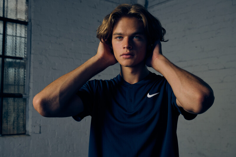 Young man in navy Nike t-shirt with hands behind head, posed against industrial white brick wall with window.