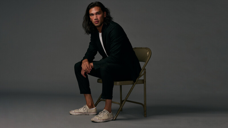 Man in black blazer and canvas sneakers sitting on metal folding chair against gray backdrop.
