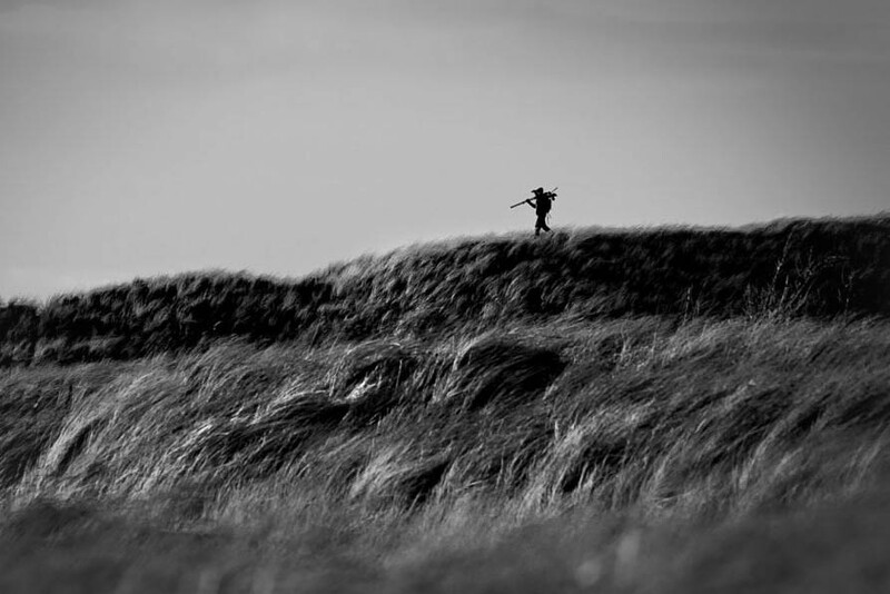 Solitary figure standing on a windswept grass-covered dune under overcast sky.