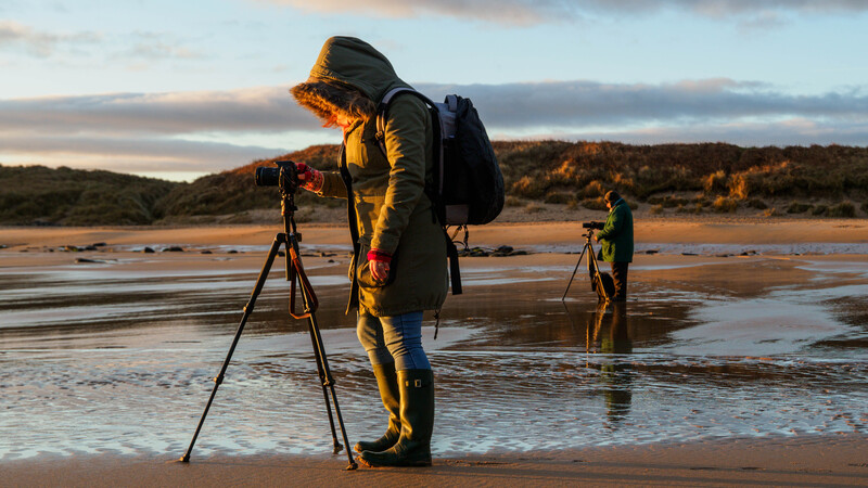 Photographer in yellow jacket adjusting tripod-mounted camera on wet beach at golden hour.