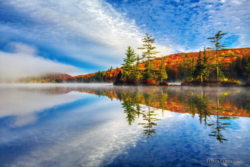 Calm lake reflecting autumn forest and mountains under blue sky with morning mist.