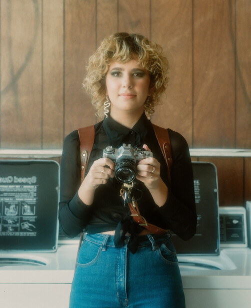 Photographer holding a vintage film camera against their chest, wearing dark jacket and blue denim skirt.
