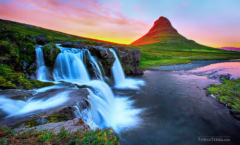 Cascading waterfalls flowing into a river with a distinctive cone-shaped red mountain rising beyond at sunset.