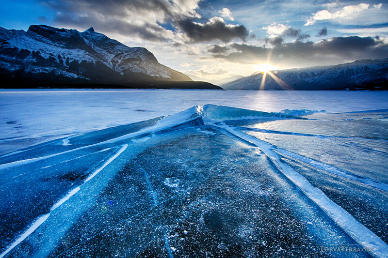 Frozen lake with blue ice cracks stretching toward snow-capped mountains and dramatic sunset sky.