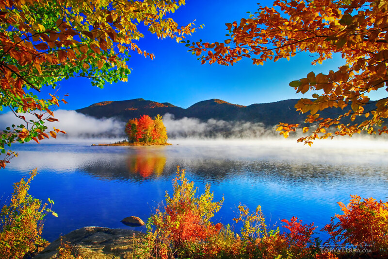 Mountain lake surrounded by vibrant autumn foliage with mist rising from still blue water.