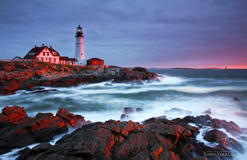 Lighthouse on rocky coastal outcrop at sunset with crashing waves and pink-purple sky.