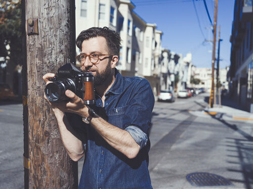 Photographer in denim shirt and glasses holding vintage film camera against weathered wooden pole on urban street.
