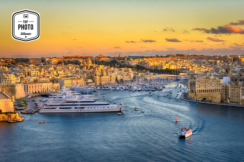 Aerial view of a Mediterranean harbor at golden hour with luxury yachts moored in blue water and warm-lit waterfront buildings.