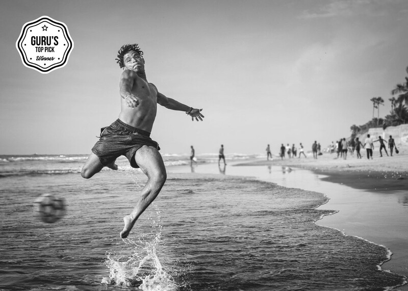Shirtless man mid-jump in shallow ocean water with beach and distant figures in background.