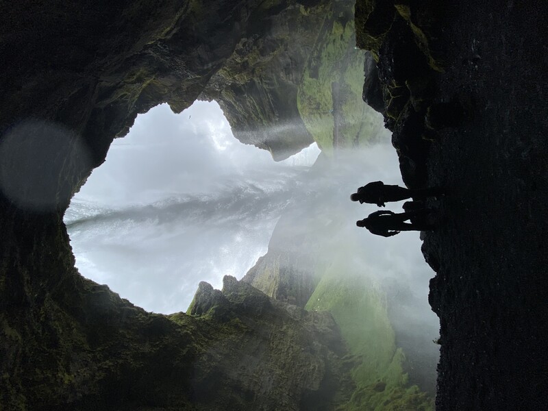 Two silhouetted figures standing in a cave opening with a waterfall cascading down the rocky cliff face above.