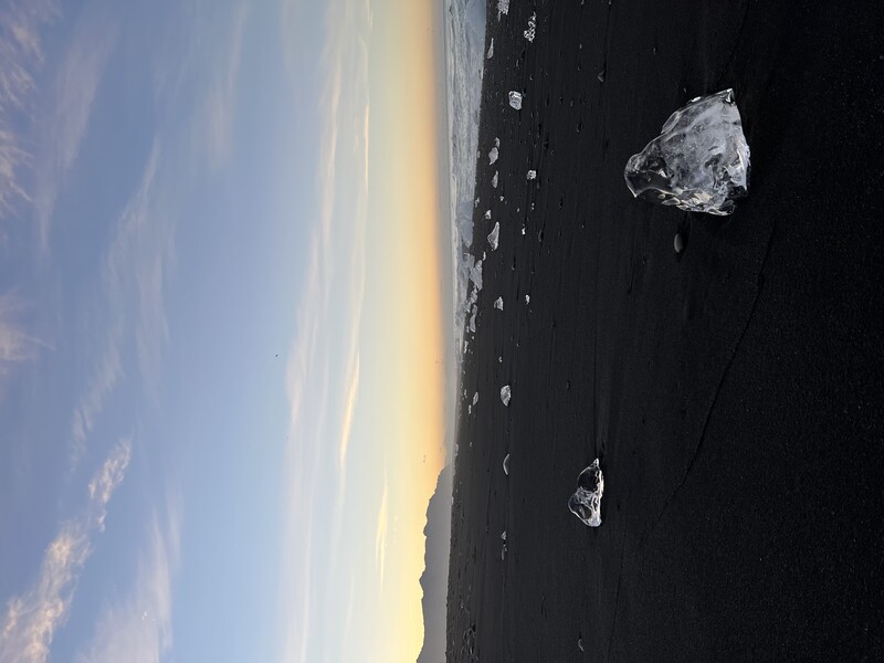 Translucent chunks of glacial ice scattered across a black sand beach at dawn, with mountains and ocean visible in the distance.