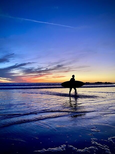 Silhouetted surfer standing in shallow water at sunset with blue and orange sky reflecting on wet sand.