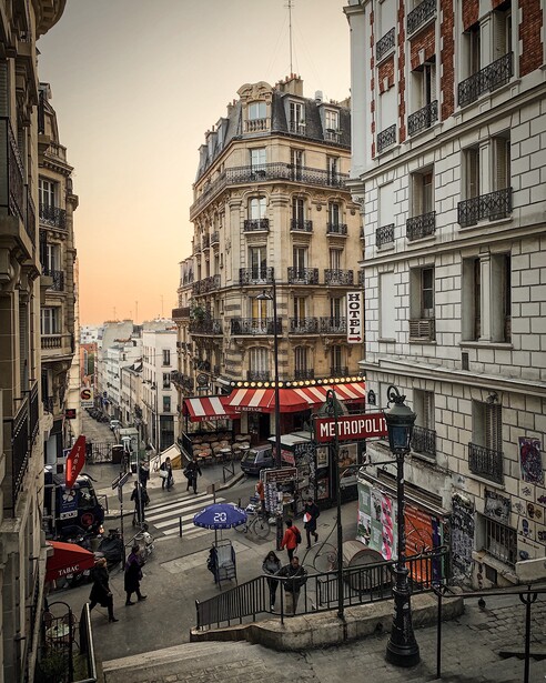 Parisian street intersection with Haussmann-style buildings and a red-awninged café at golden hour.