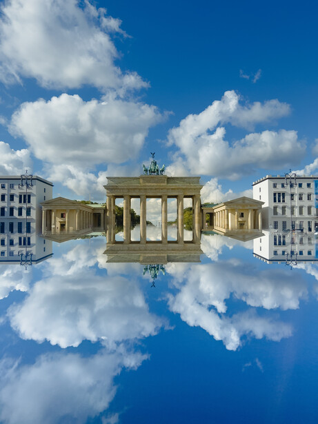 Brandenburg Gate reflected in still water against blue sky with white clouds.