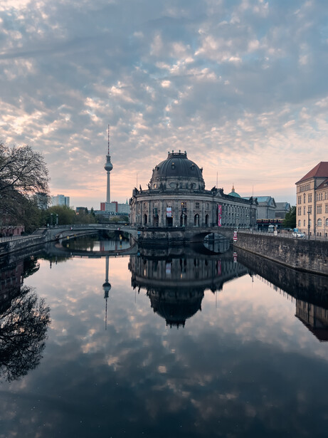 Neoclassical museum building reflected in still water at dawn, with TV tower in background.