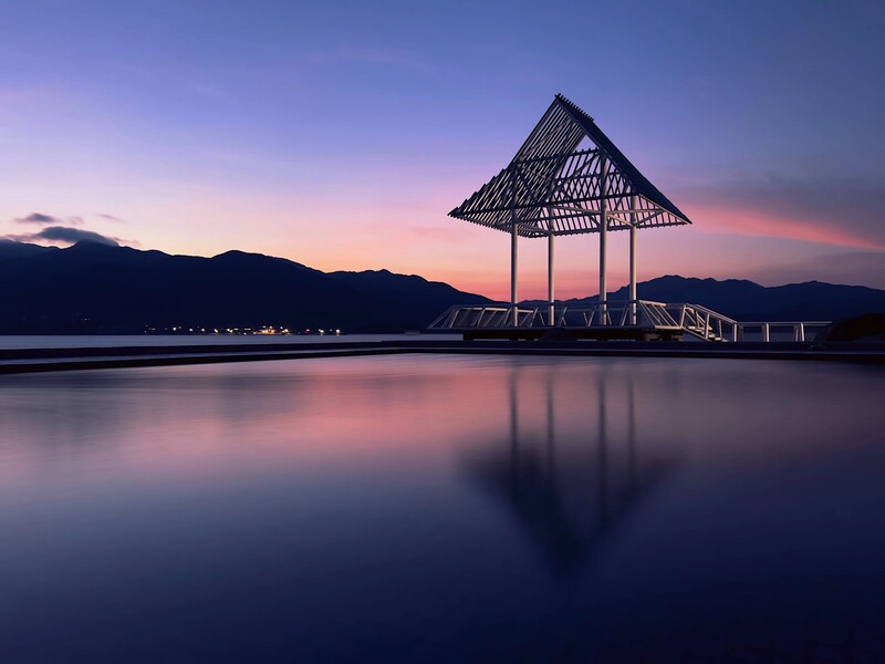 Modern pavilion silhouetted against a purple and pink sunset sky, reflected in still water with mountains in the background.