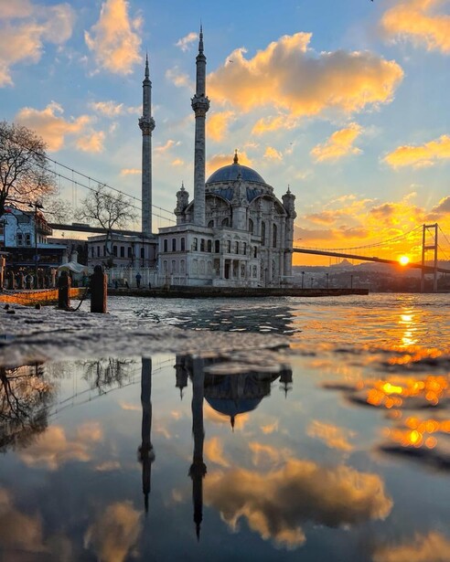 White Ottoman mosque with twin minarets reflected in still water at golden hour sunset.