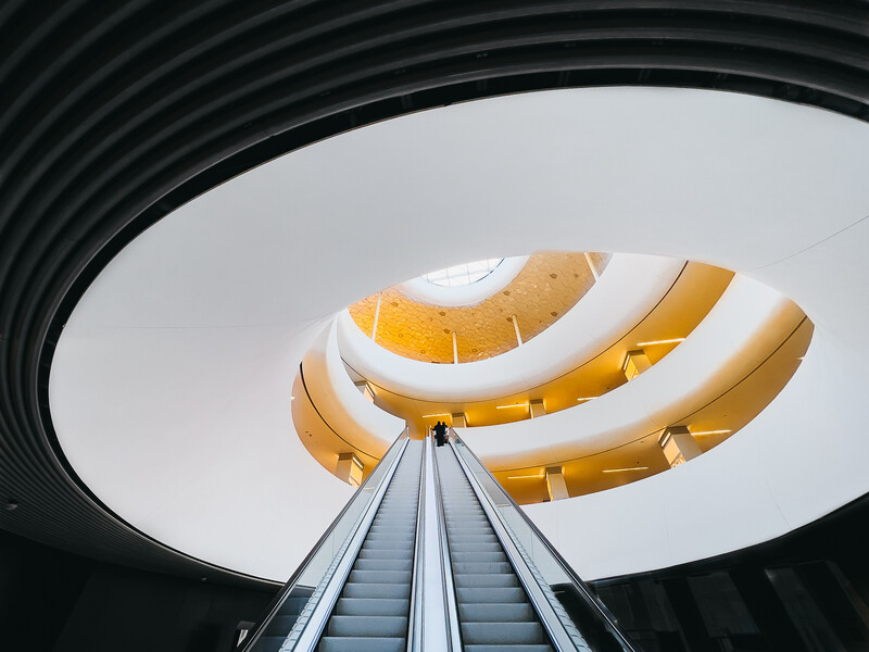 Upward view through a spiraling concrete structure with golden interior surfaces and a centered escalator.
