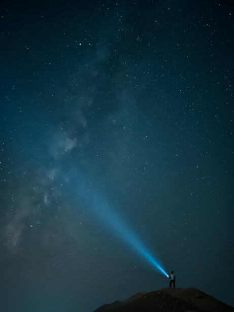 Silhouetted figure on a hilltop shining a flashlight beam toward a starry night sky with visible Milky Way.
