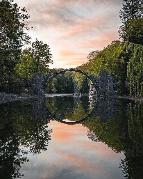 Stone arch bridge reflecting perfectly in still water at golden hour, surrounded by dense green foliage.
