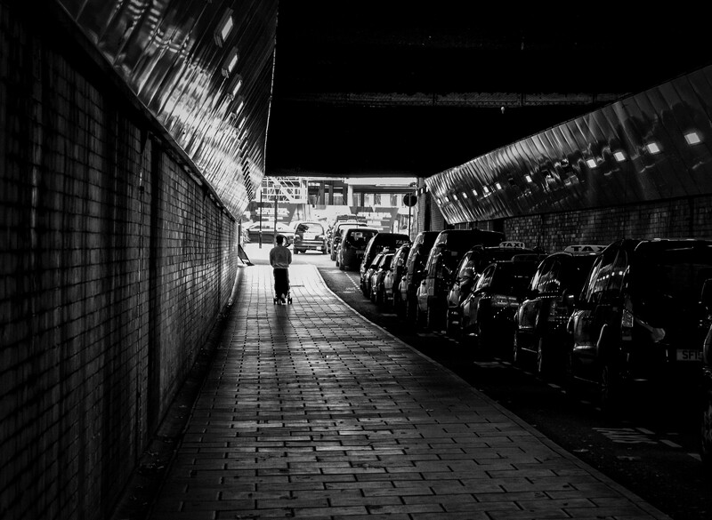 Solitary figure walking through narrow covered passage lined with parked cars and brick walls.