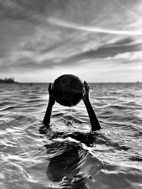 Person emerging from ocean water with head bowed and arms outstretched, captured in black and white.