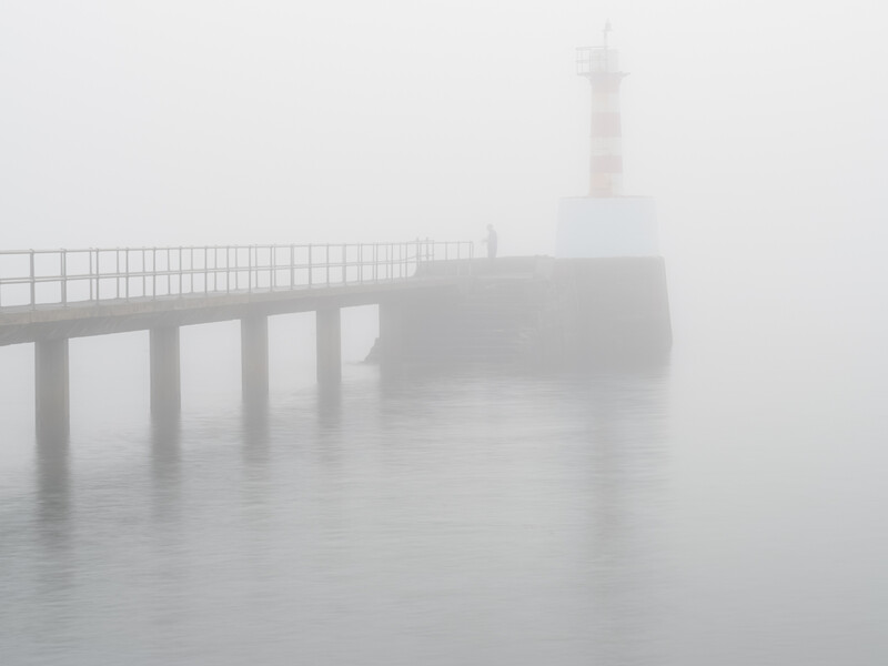 Concrete pier and lighthouse silhouette emerging through dense fog over calm water.